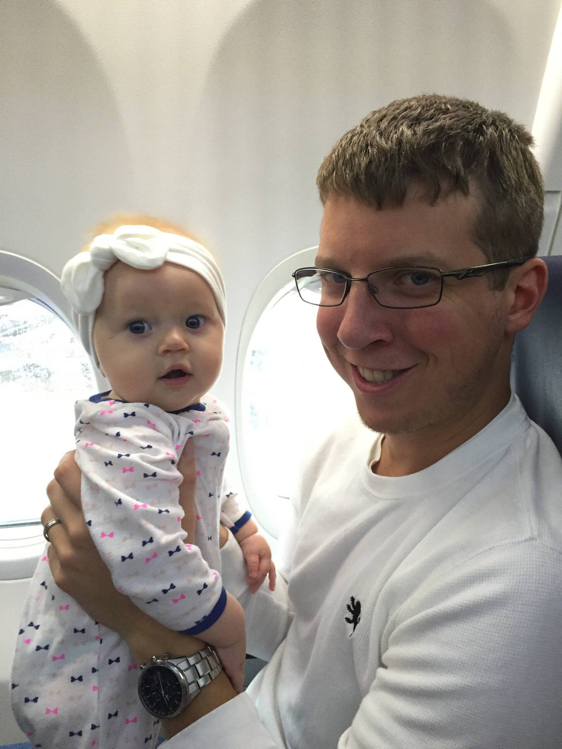 Man in glasses holding baby with white bow headband and patterned onesie while seated by an airplane window.