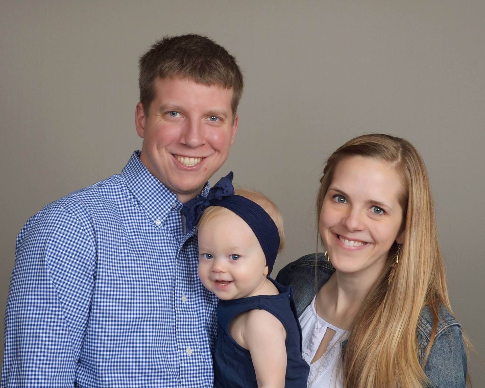 Smiling couple holding a baby in a navy headband; man in blue checked shirt, woman with long blonde hair and denim jacket.
