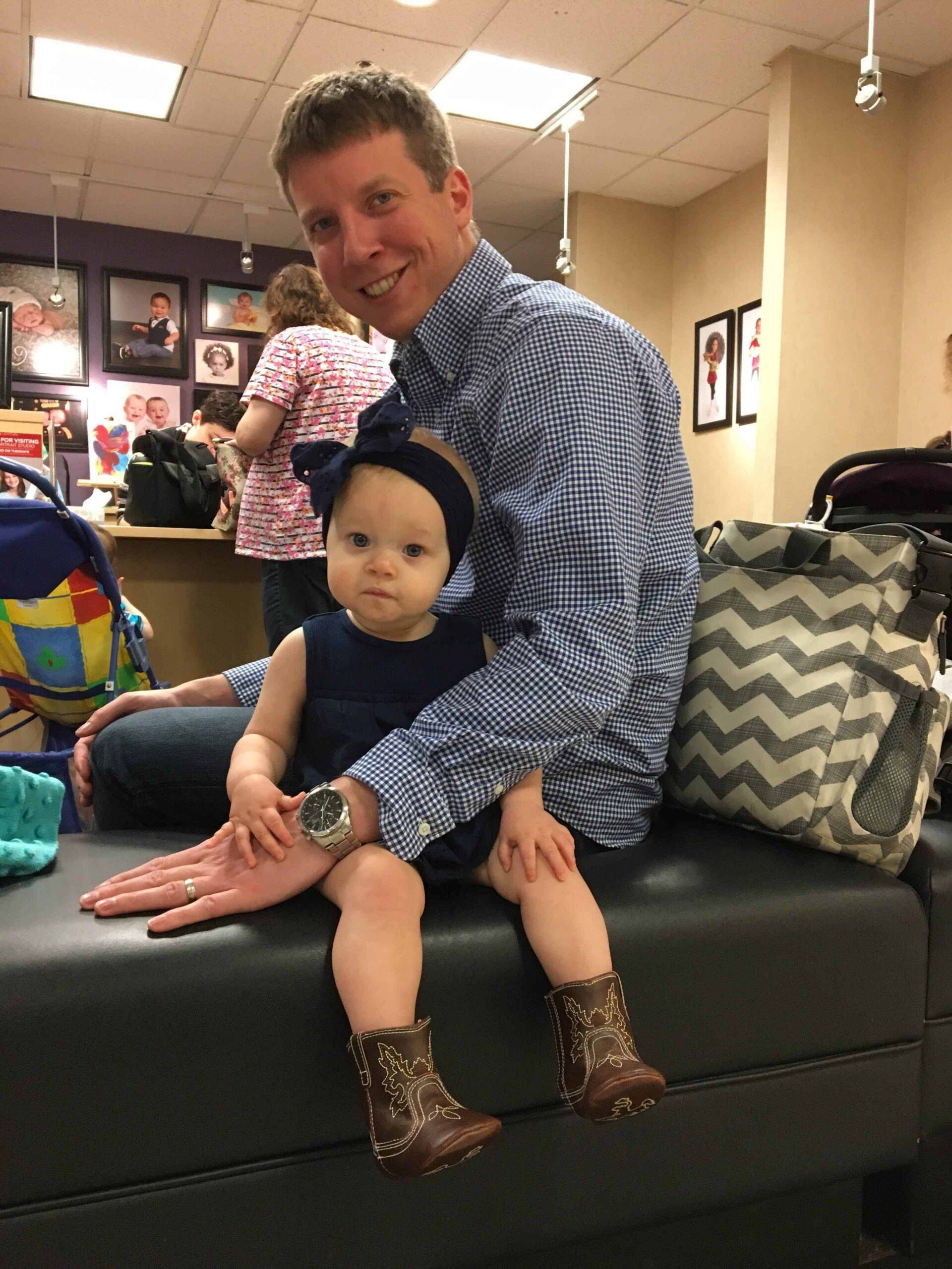 Man in blue checked shirt holding toddler wearing navy headband and brown cowboy boots, seated on a studio bench.