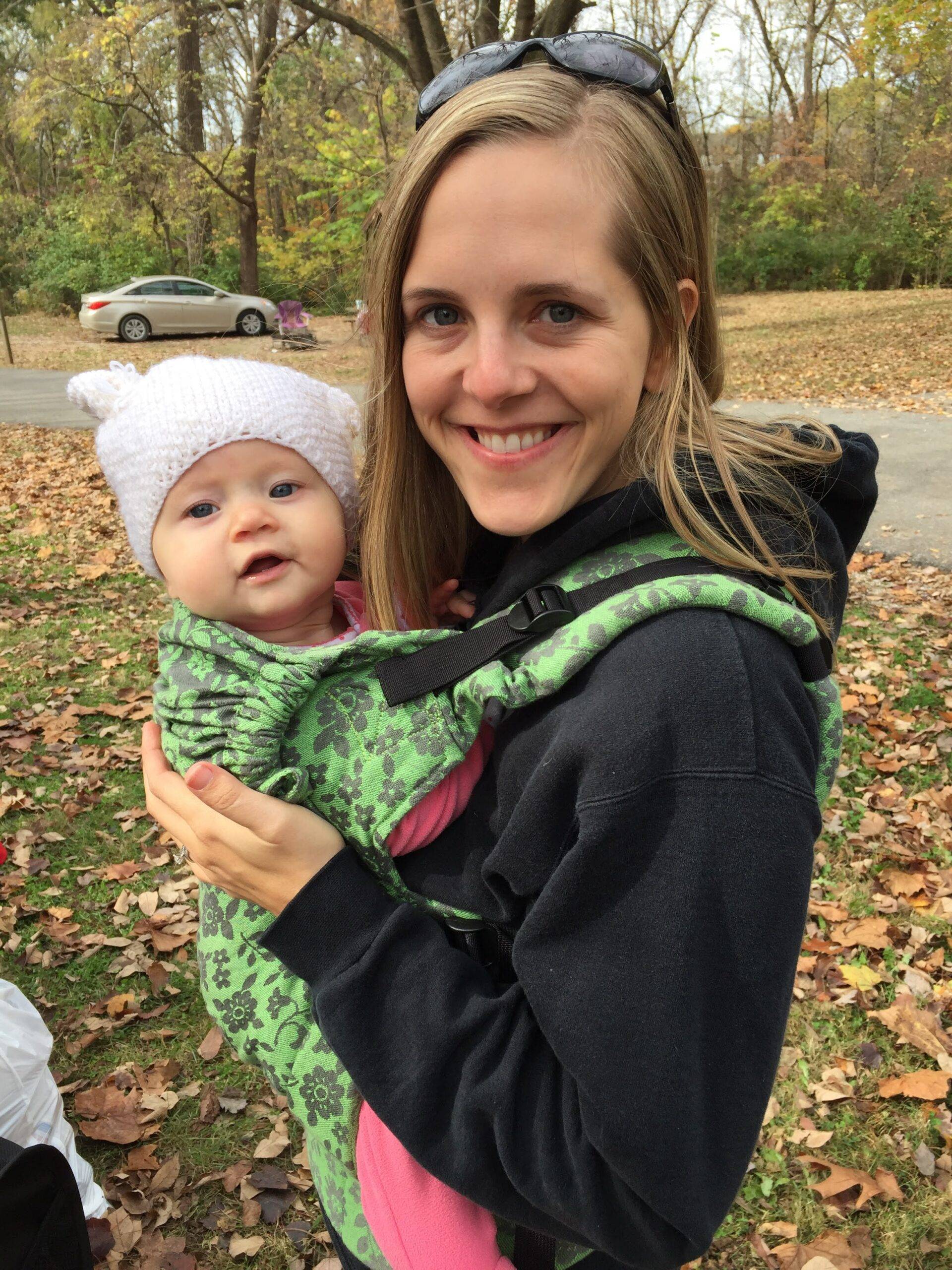 Smiling woman holding a baby in a green floral carrier; baby wearing a white knit hat in a park with fallen leaves.