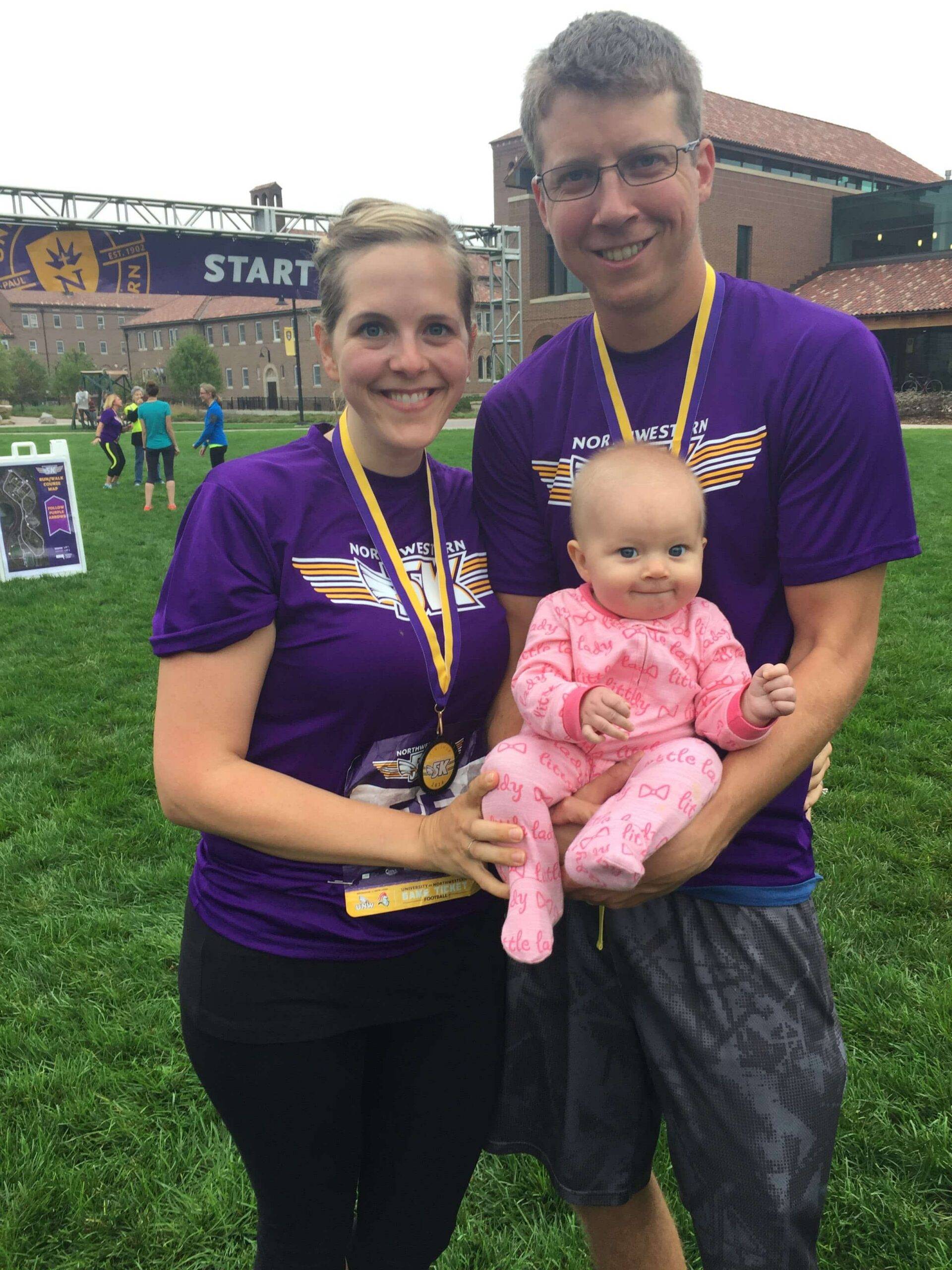 Smiling couple in matching purple race shirts and medals holding a baby in a pink onesie on a grassy field near START banner.