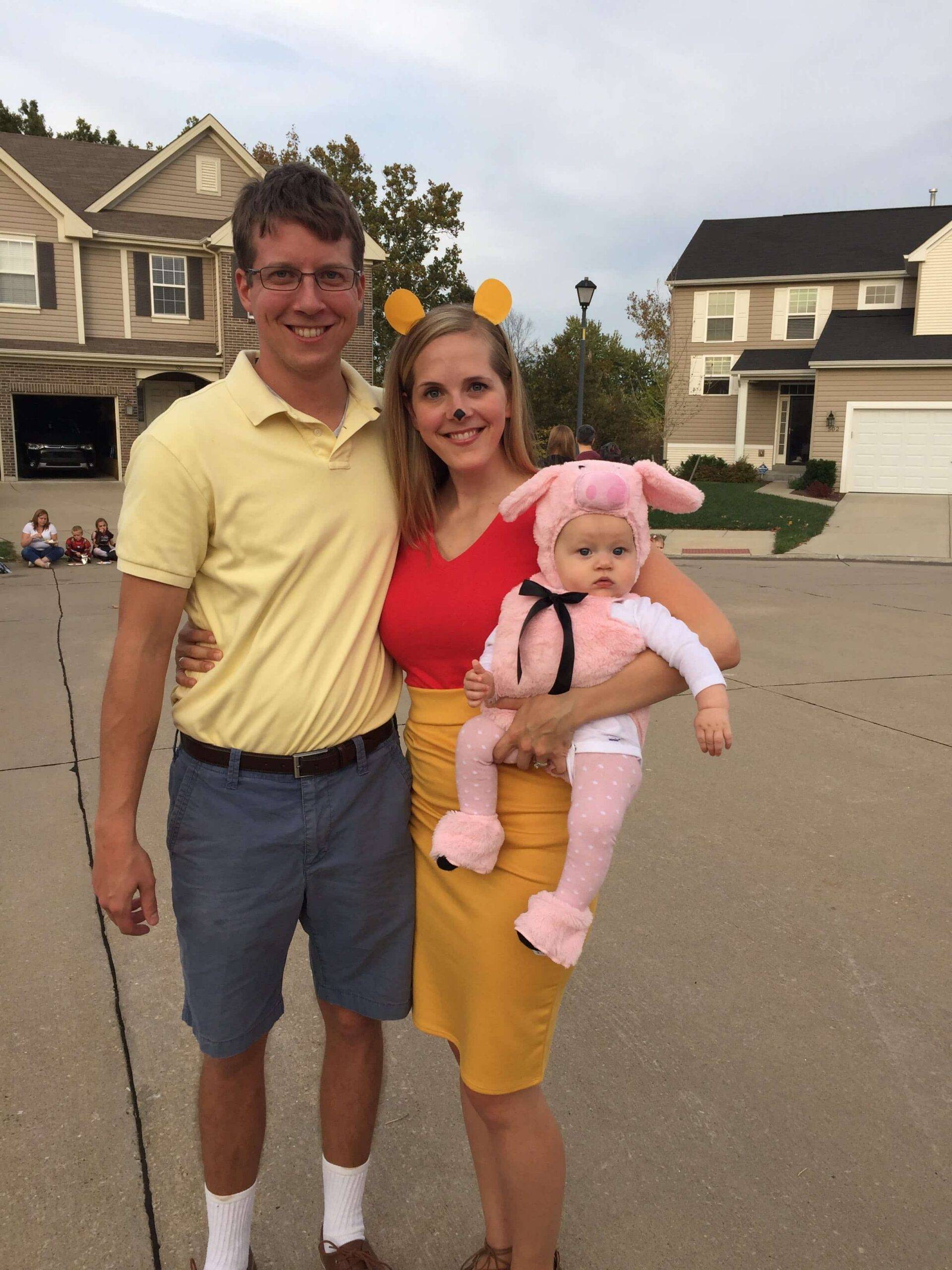 Family on a suburban street: man in yellow polo, woman with bear ears in red and yellow, baby in a pink pig costume.