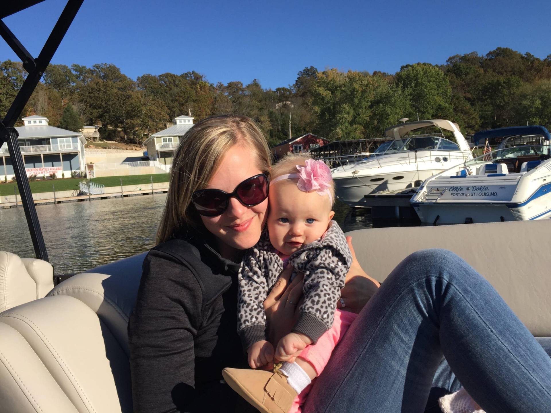 Woman wearing sunglasses holding a baby with a pink flower headband while seated on a boat with a marina in the background.