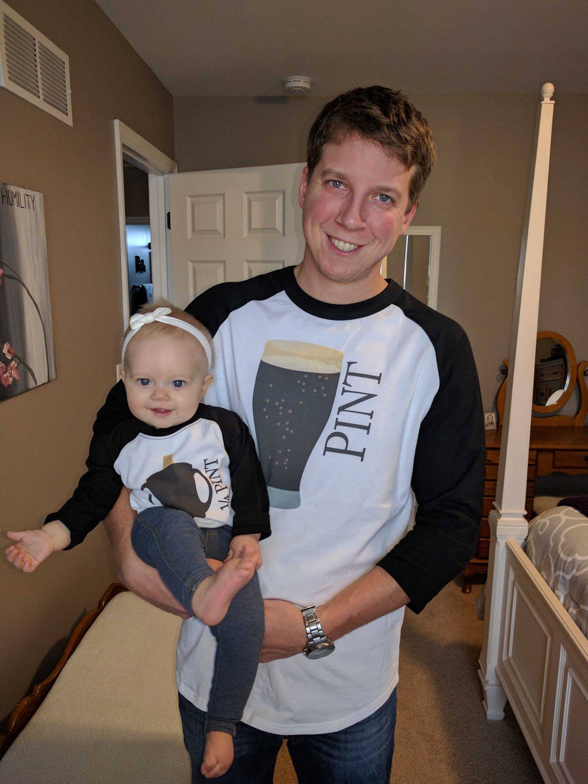 Smiling man holding a barefoot baby with a white headband; both wear matching baseball shirts with a pint-glass graphic.