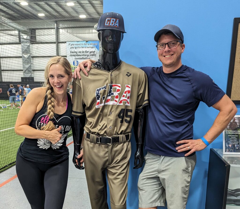 Two smiling adults pose with a black mannequin wearing a GBA baseball uniform (No. 45) inside an indoor sports facility.