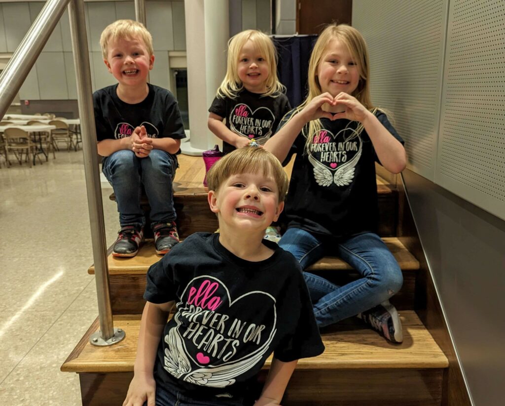 Four smiling children in matching black Ella Forever In Our Hearts shirts sitting on wooden steps, one forming a heart with her hands.