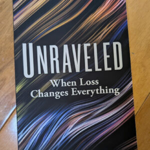 Book cover reading UNRAVELED — When Loss Changes Everything, colorful flowing strands on black background, on a wooden floor