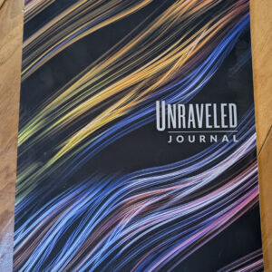Black journal titled "Unraveled Journal" with multicolored, thread-like flowing streaks across a black cover, resting on a wooden floor.