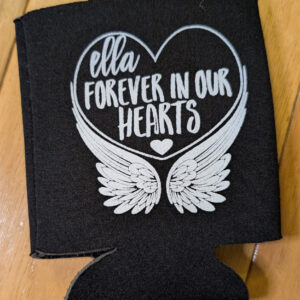 Black can cooler with white heart, angel wings and text reading Ella forever in our hearts on a wooden table.