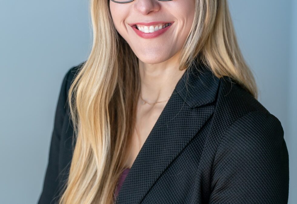 Smiling woman with long blonde hair and black glasses wearing a black blazer against a light gray background.