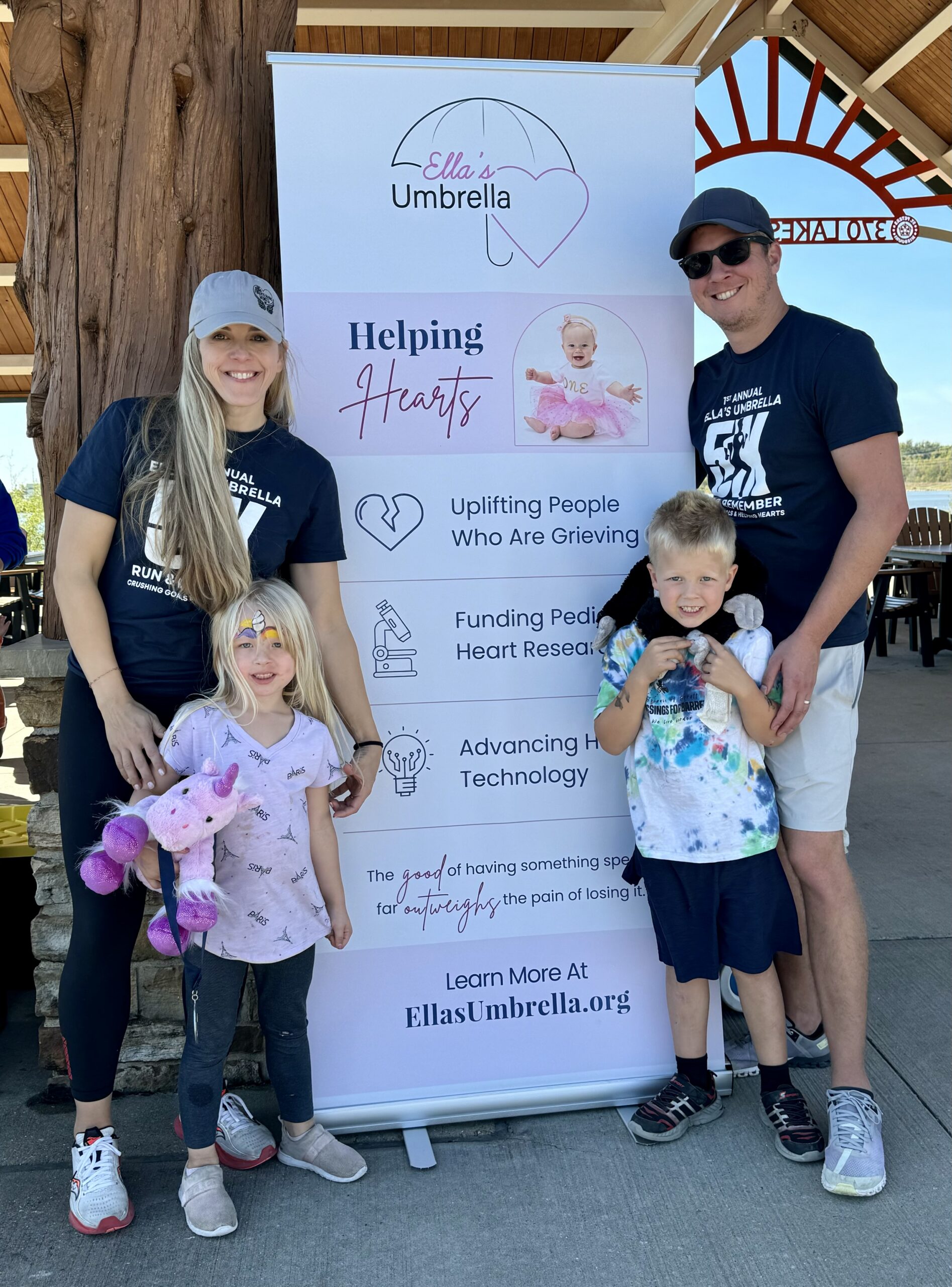 Family of four beside Ella's Umbrella banner reading "Helping Hearts"; little girl holding a purple unicorn plush.