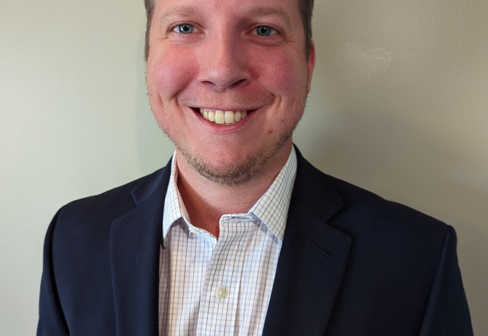 Smiling man with fair complexion and short brown hair wearing a navy blazer and checked shirt against a beige wall