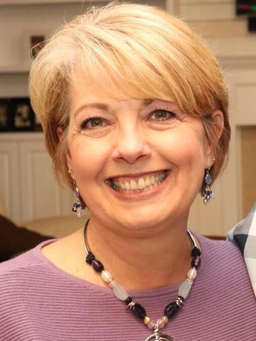 Smiling woman with short blonde hair wearing a purple top and beaded necklace with round pendant, indoor setting