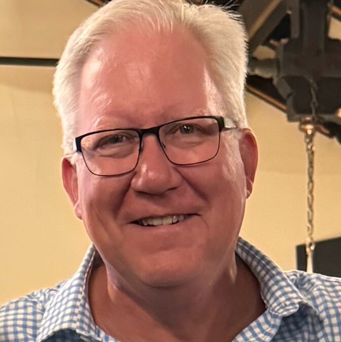 Smiling middle-aged man with short white hair and glasses wearing a blue checkered shirt, indoor setting