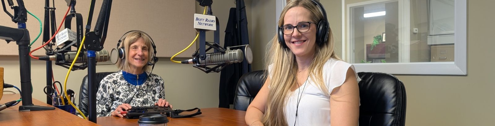 Two women wearing headphones smiling at a radio studio table with microphones labeled Bott Radio Network.
