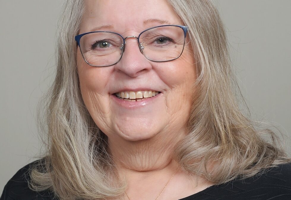 Smiling older woman with gray hair, blue-rimmed glasses, a gold heart pendant, and a black top.