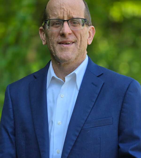 Man with glasses in a navy suit and white shirt standing outdoors with blurred green foliage in the background.