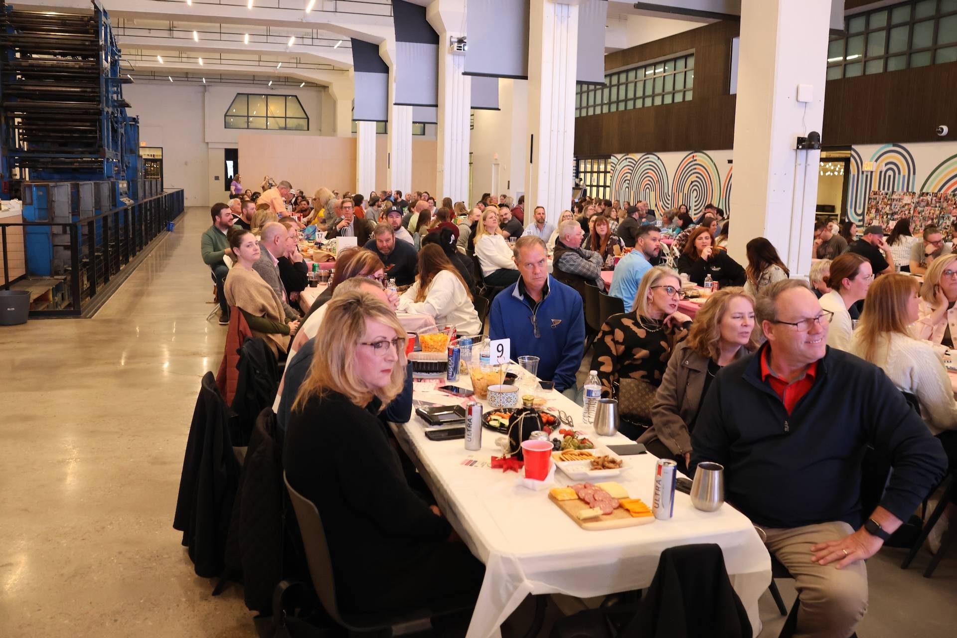 Large group of adults seated at long tables with food and drinks in a spacious industrial-style hall.