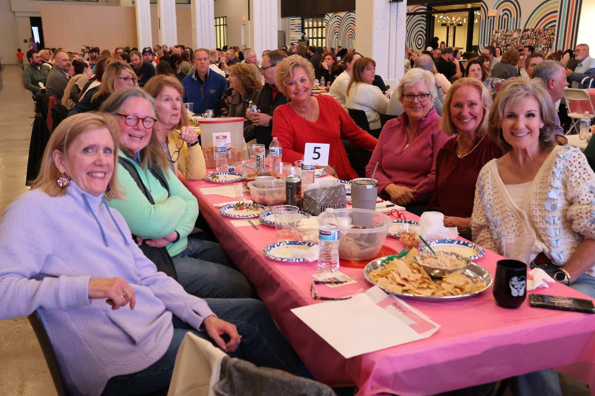 Eight women smiling at the camera around a pink-covered banquet table with plates, chips, drinks, and table number 5 in a busy hall.