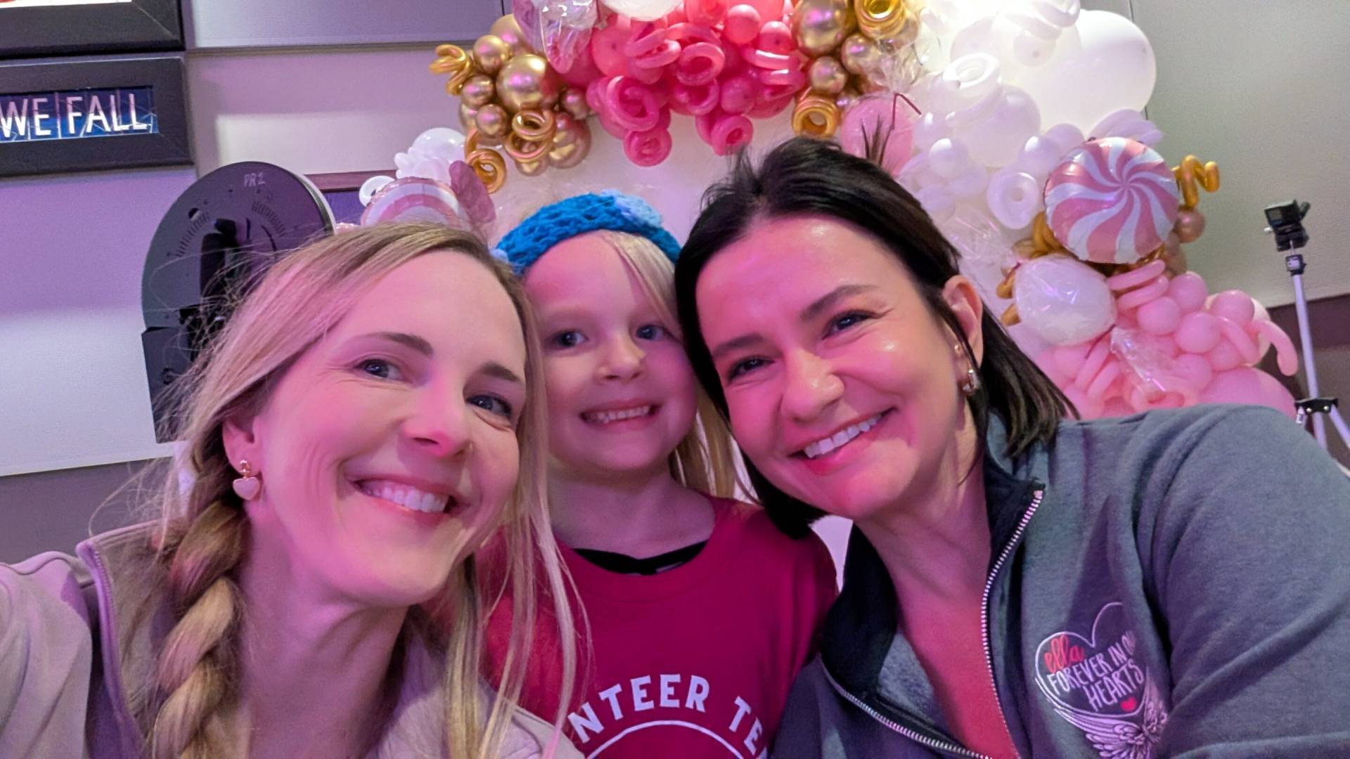 Three smiling people (two adult women and a young girl wearing a blue knit headband) posing closely in front of a pink and gold balloon arch.