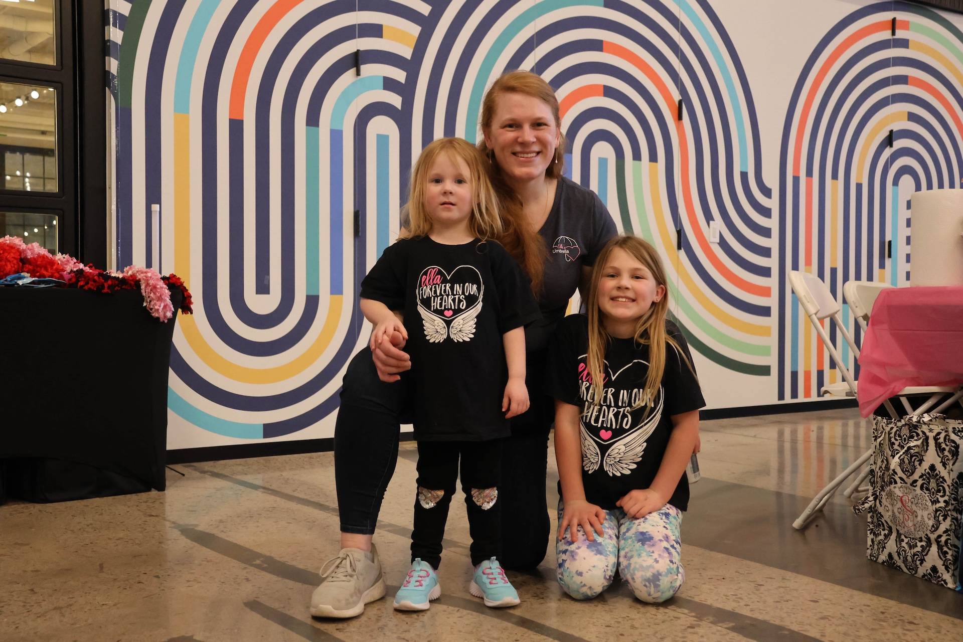 Woman kneeling with two girls in matching black shirts reading Forever in Our Hearts, colorful arched mural behind.