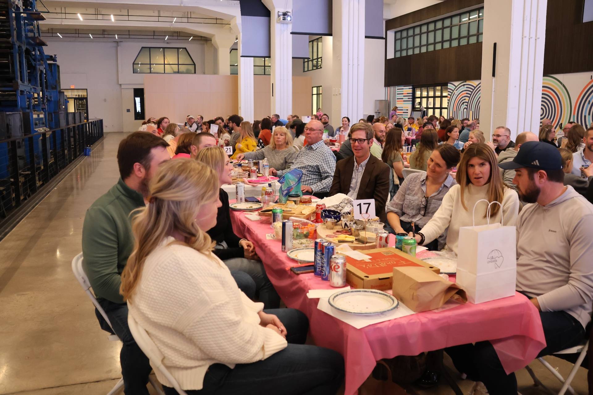 Crowd seated at long pink-covered tables eating and socializing in a large indoor hall, table number 17 visible.