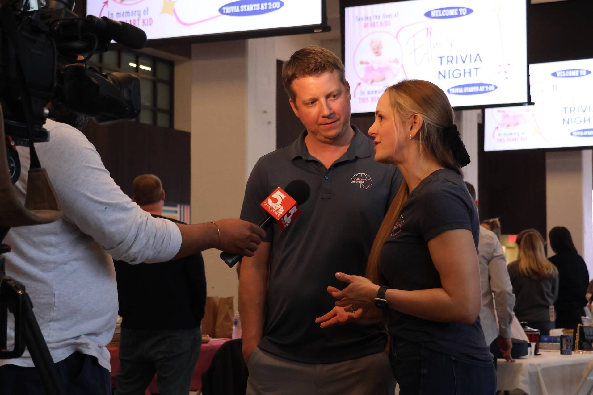 Man and woman in matching dark polo shirts speaking to a TV reporter holding a red 5 microphone at an indoor trivia night.