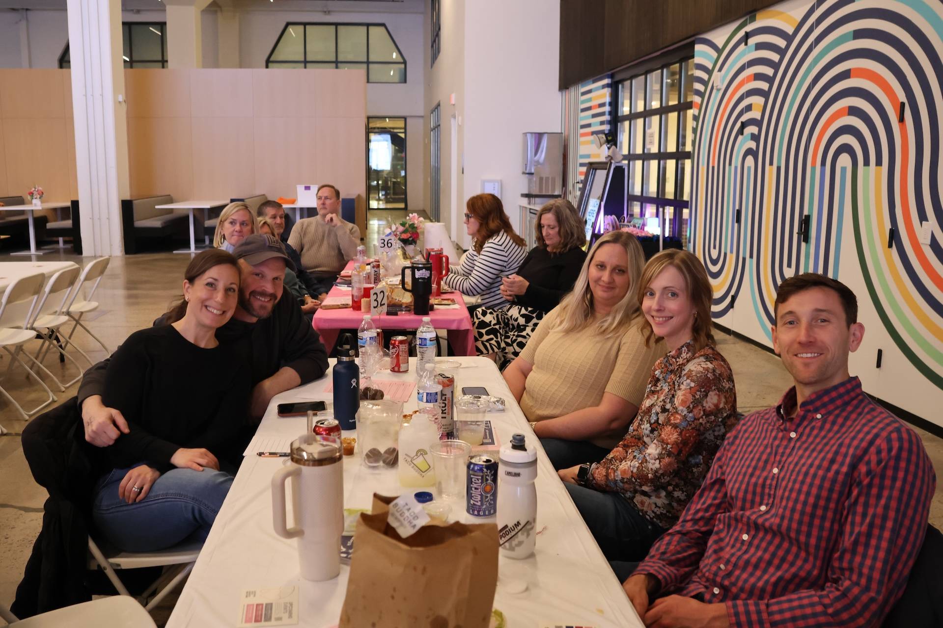 Adults seated at a long indoor table, smiling with drinks on the table and a colorful concentric wall mural behind them.