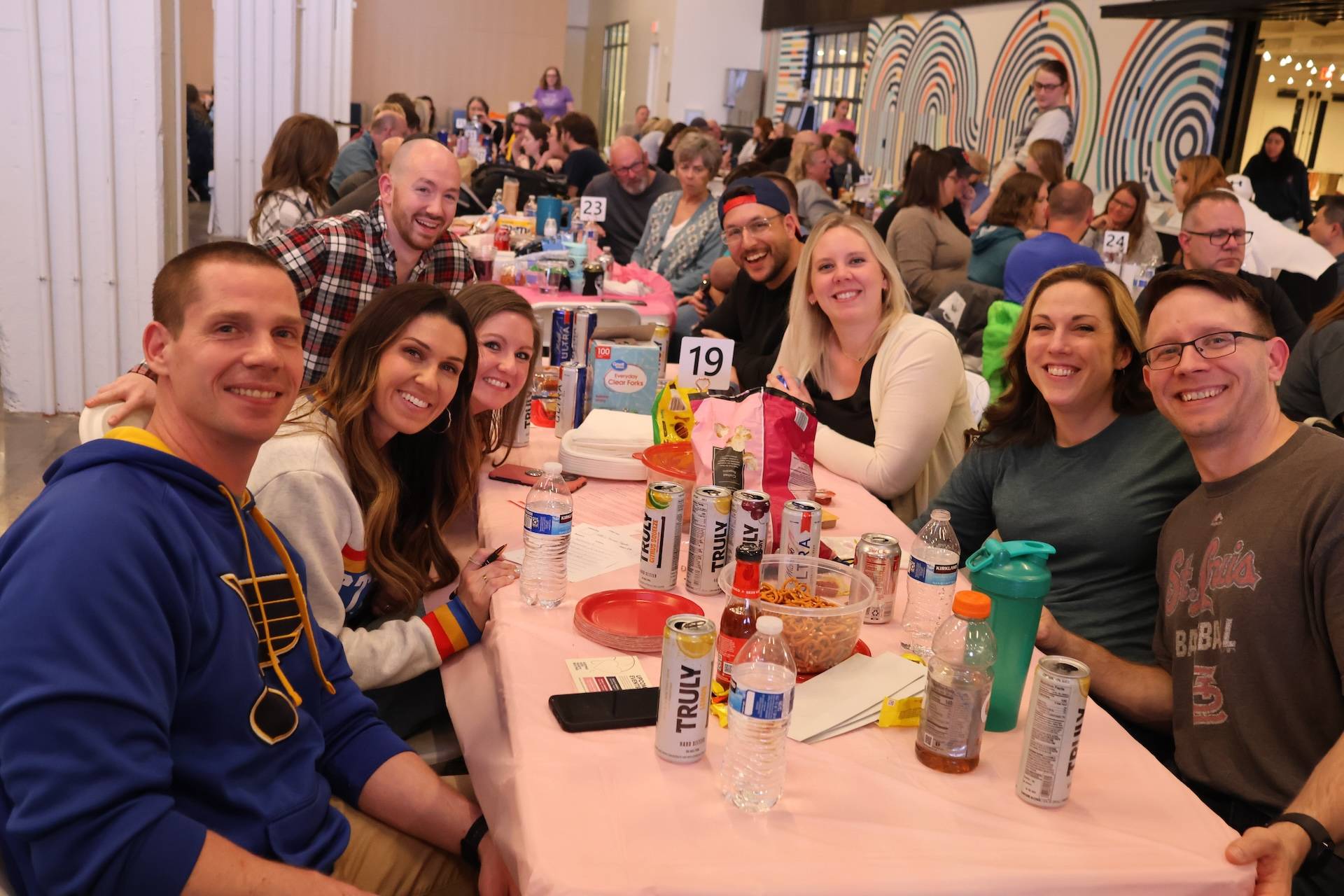 Nine smiling adults seated around a long table with drinks, snacks, and a table number 19 at an indoor gathering.