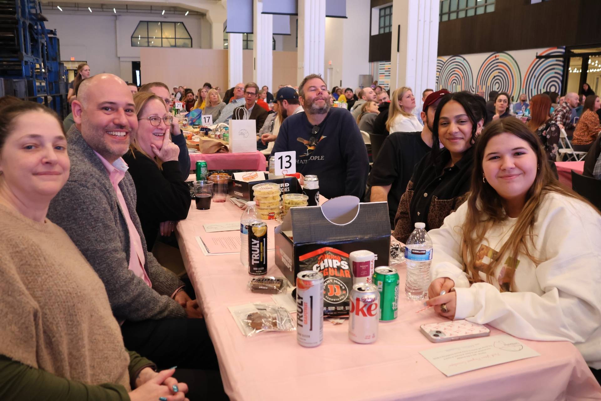Group of smiling people seated around a table with snacks, drinks and a table number 13 at an indoor event.