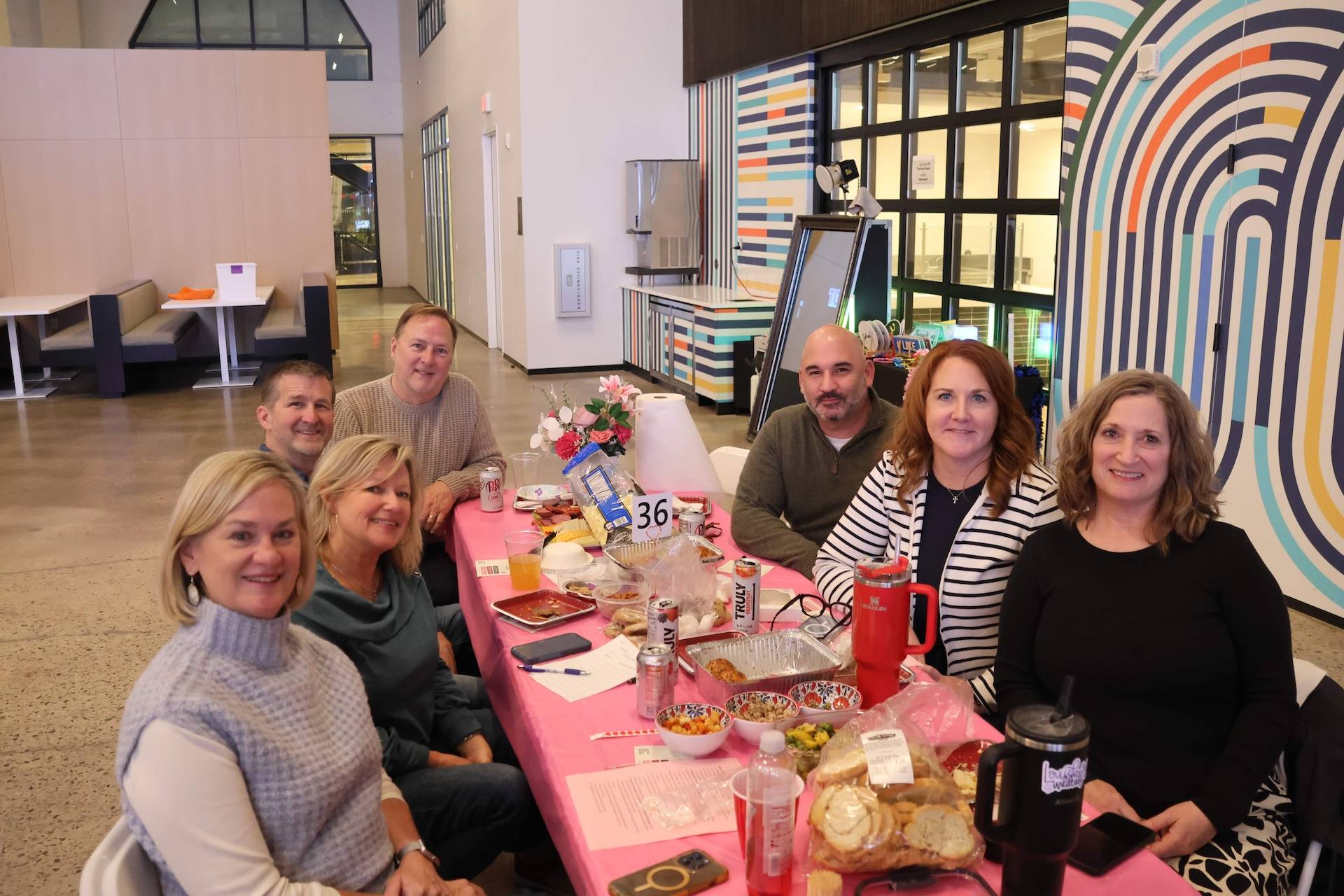 Eight people around a pink-covered table with food and drinks, smiling indoors; table number 36.