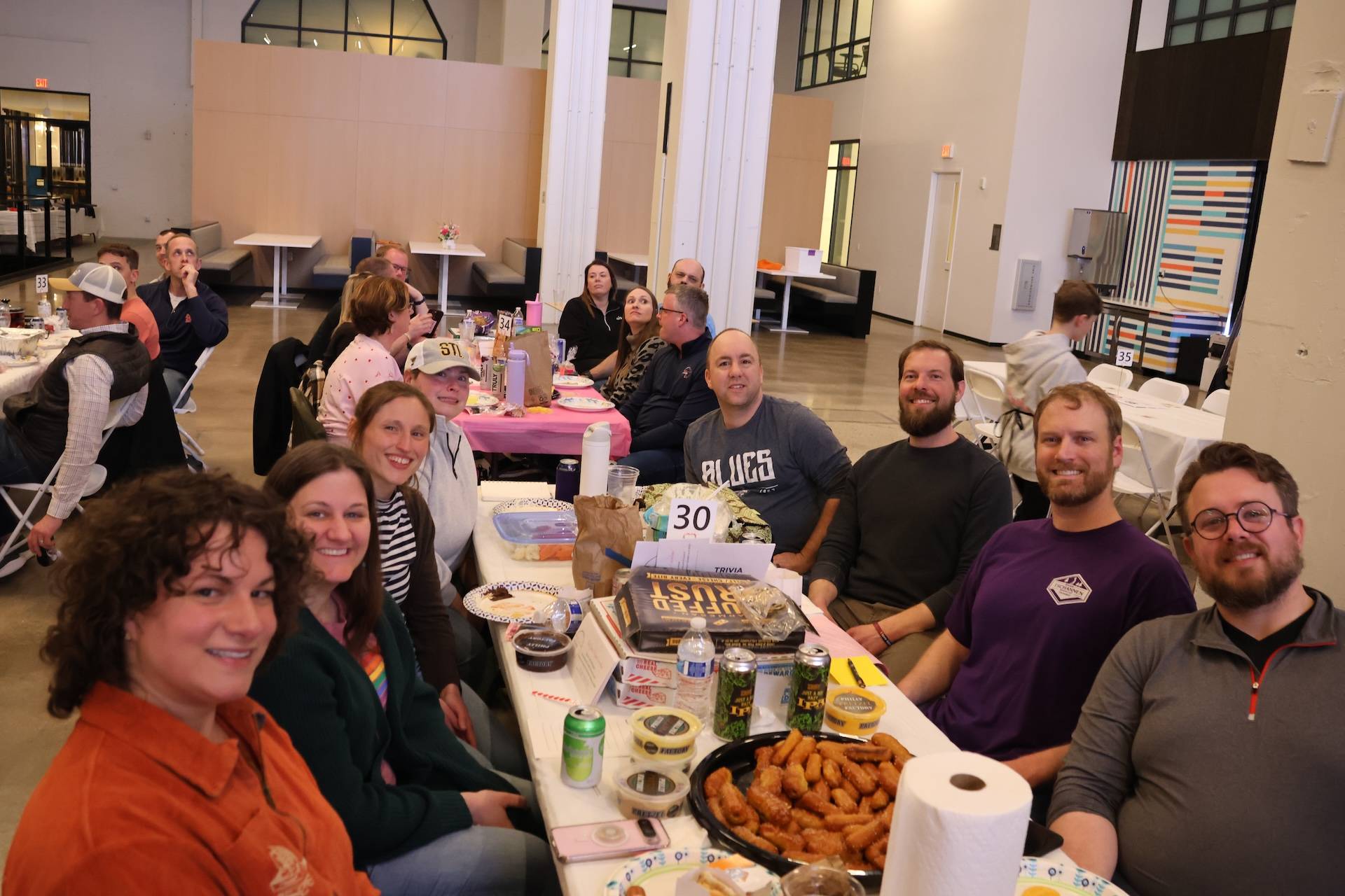 Group of smiling adults seated around a long table indoors with snacks, drinks and a table number 30 at a social gathering.