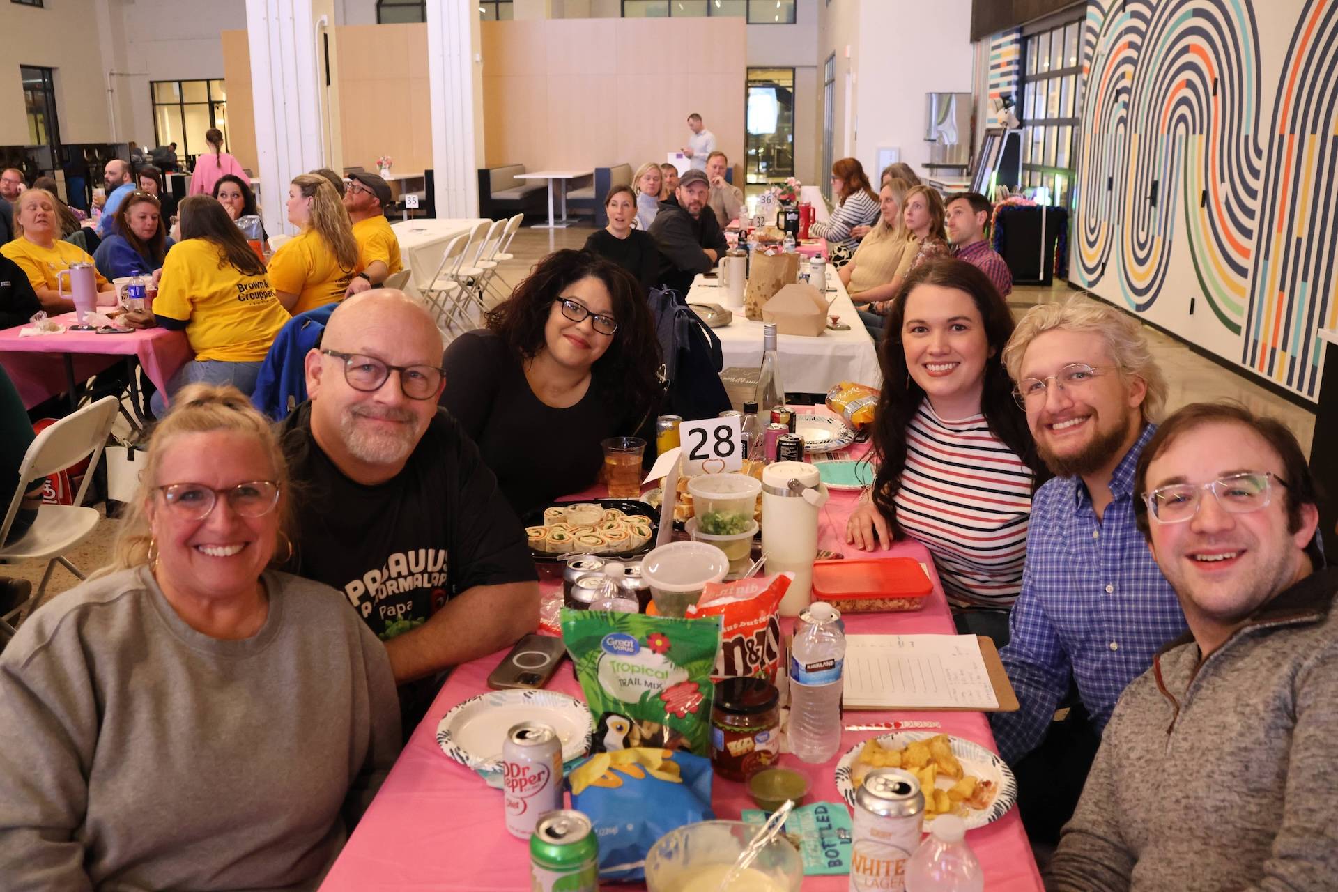 Group of seven smiling adults seated at a pink table with snacks, drinks and a table number 28 at an indoor gathering