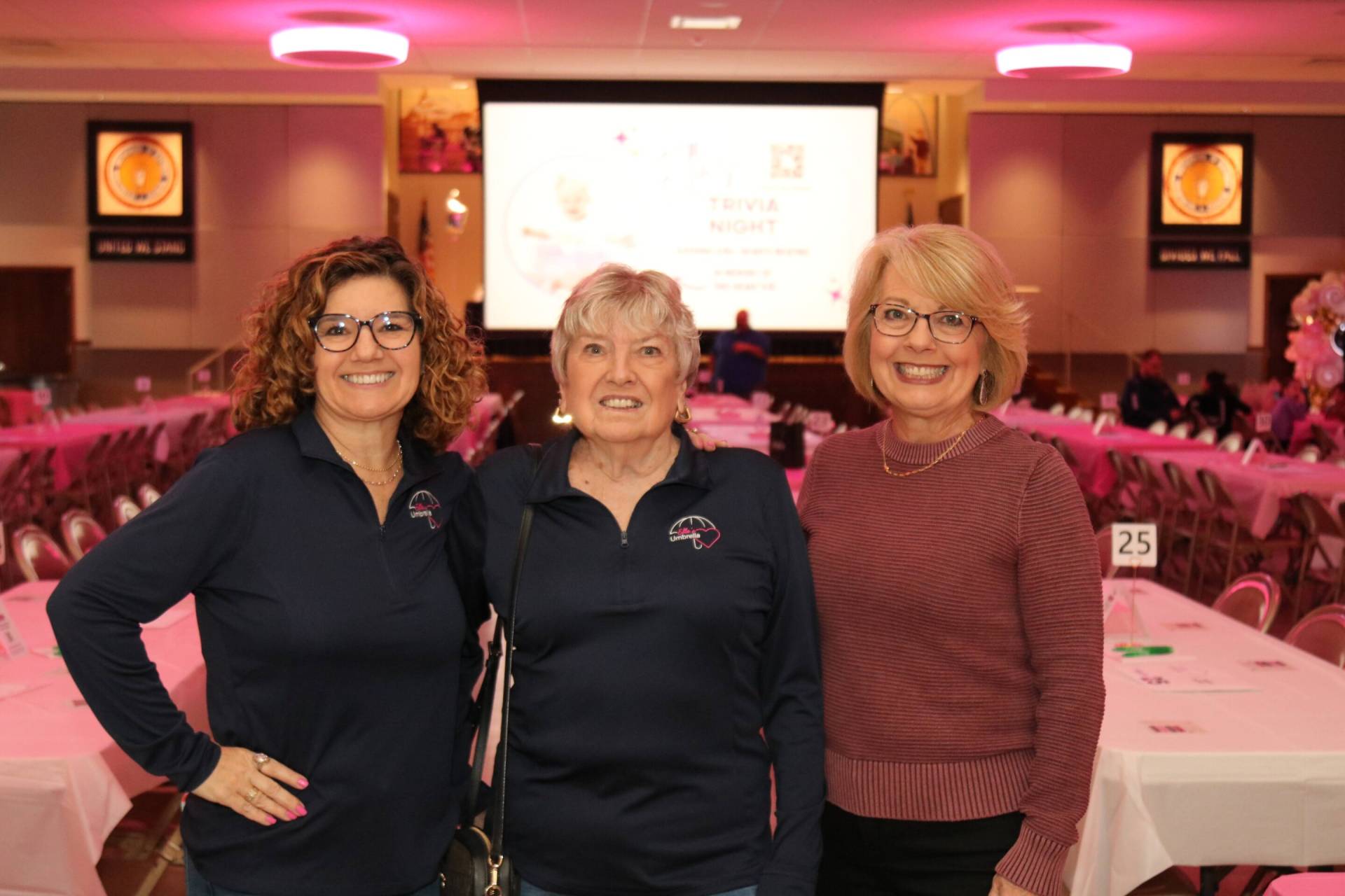 Three smiling middle-aged women standing arm-in-arm in a pink-lit banquet hall with rows of numbered tables and a projection screen behind them.