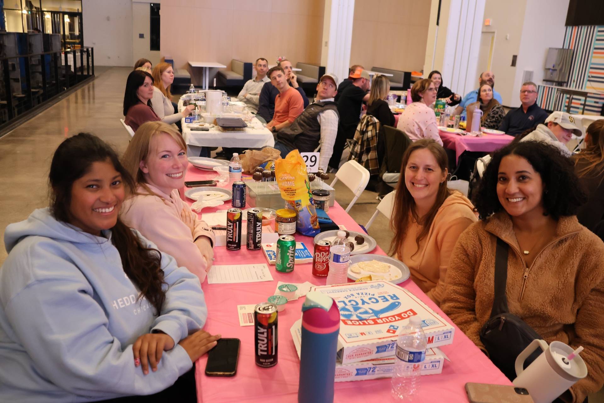 Four smiling women seated at a pink-tablecloth table with drinks, snacks and pizza boxes in a busy indoor gathering.