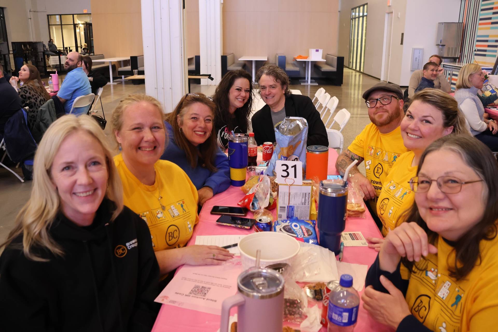 Smiling group of adults around a pink table with snacks and drinks, several wearing matching yellow BC shirts, table number 31.
