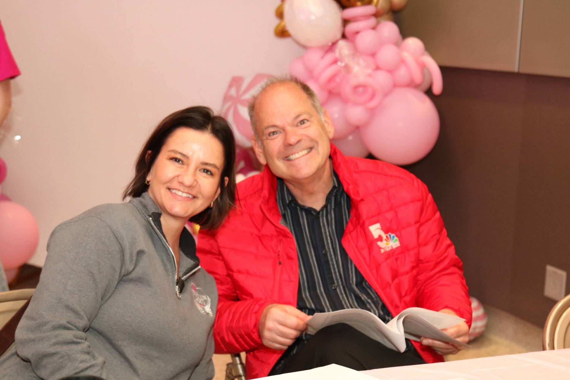 Two smiling adults seated together at a table, woman in a grey zip sweatshirt and man in a red jacket holding papers, pink balloon decor behind them