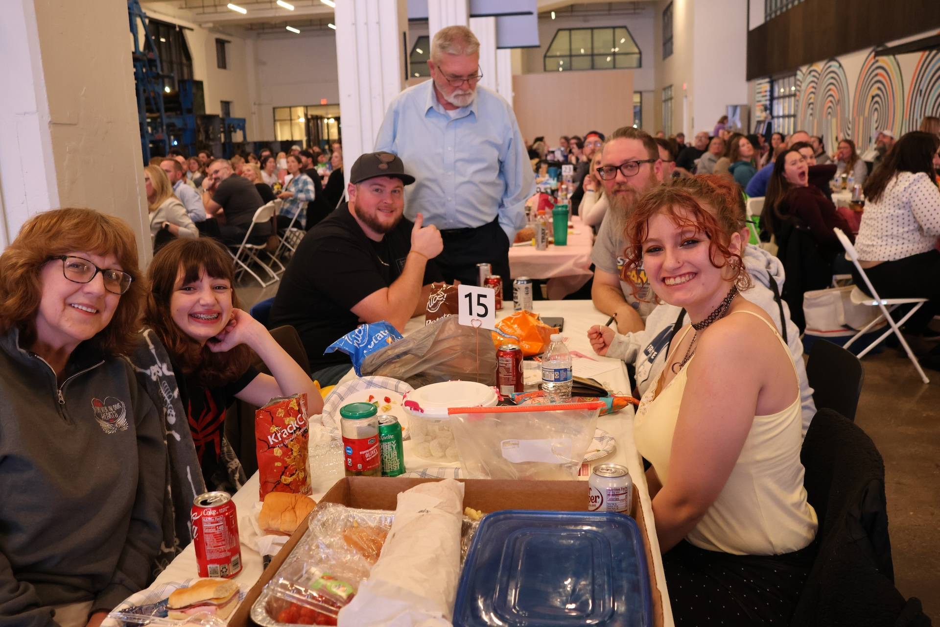 Small group of people smiling at a crowded indoor event, seated around table 15 with food.