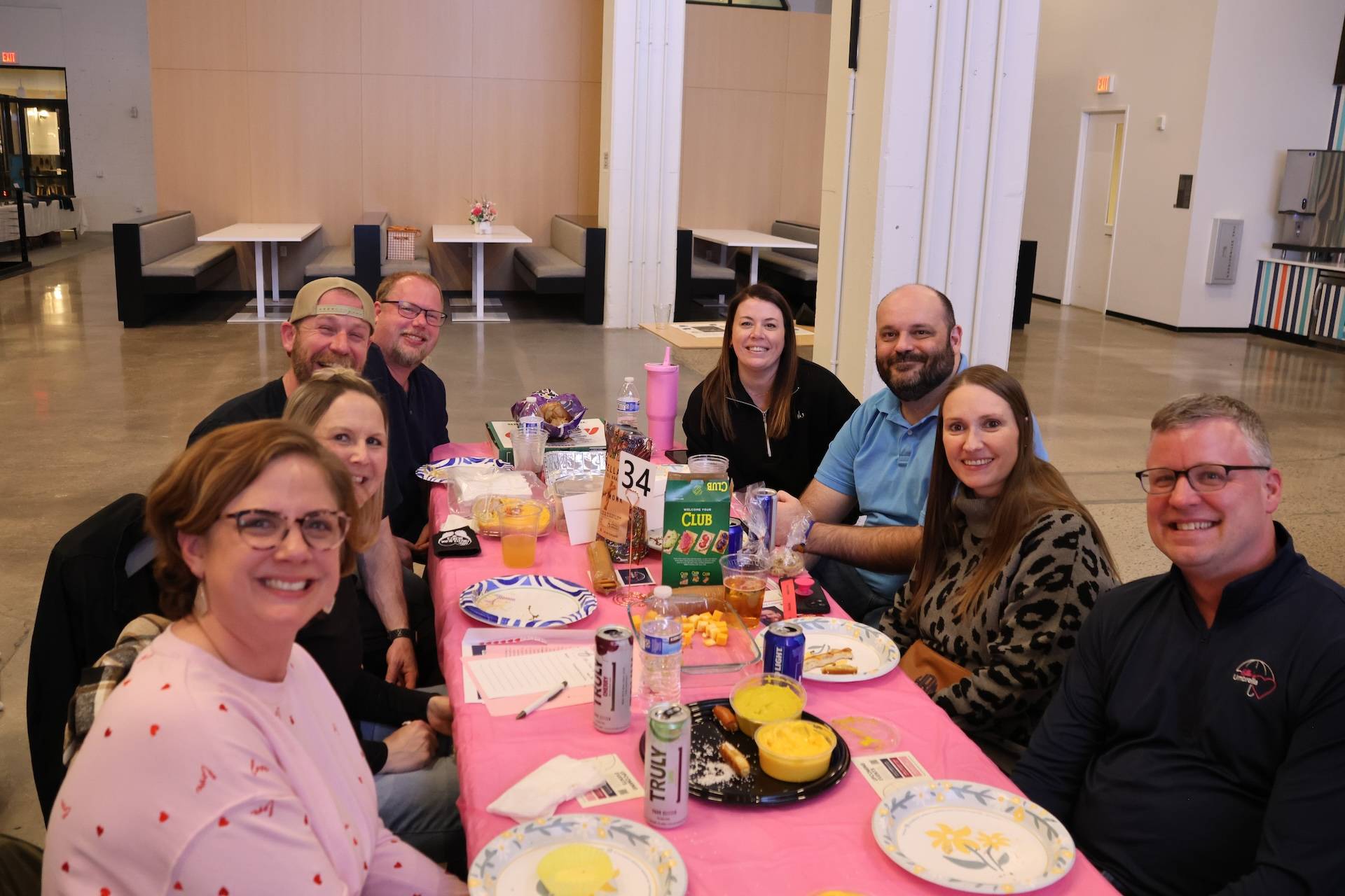 Nine adults seated around a pink-covered table with snacks and drinks, smiling at the camera in a large indoor space.