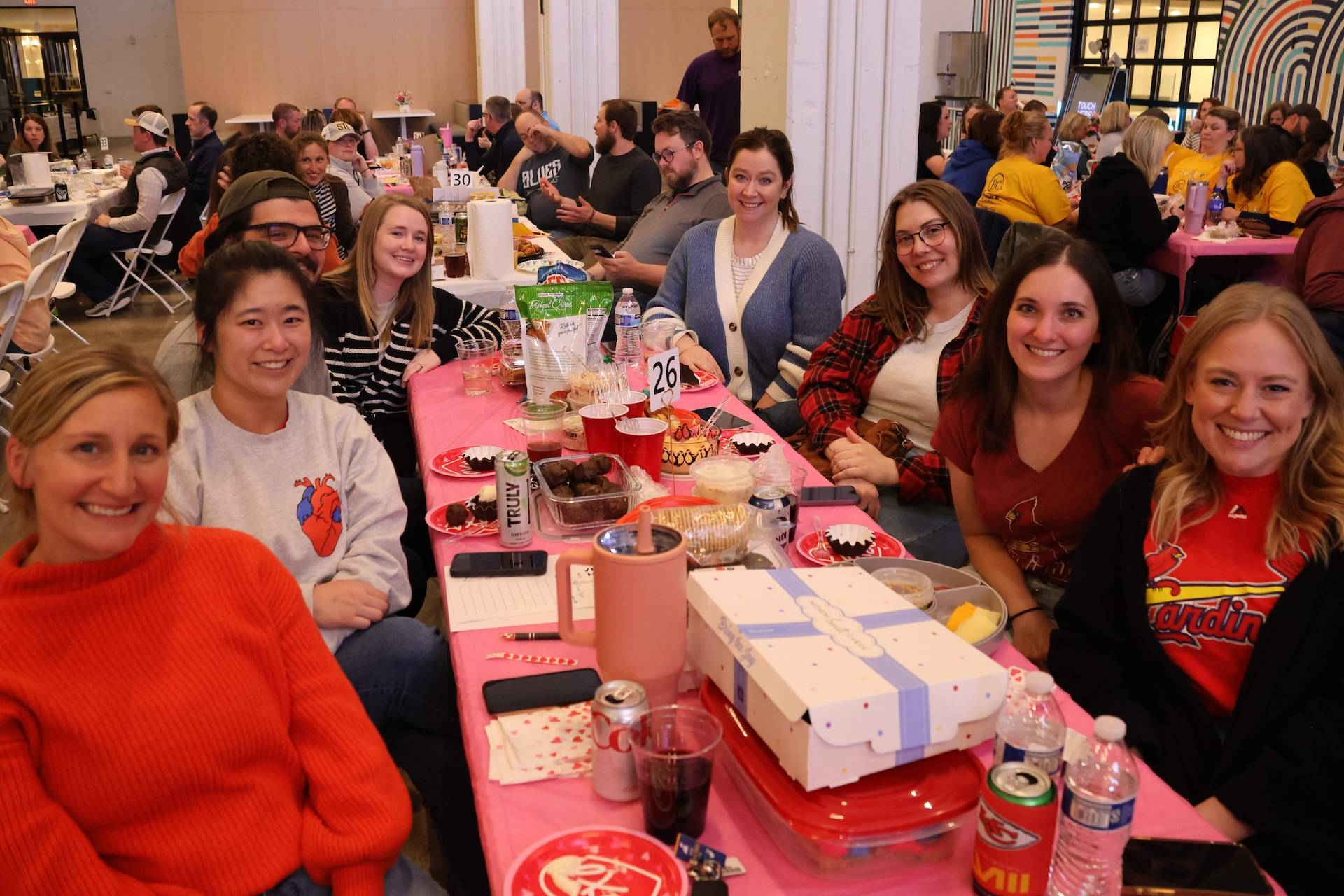 Smiling group at a pink-covered table with snacks, drinks and a visible table number 26.