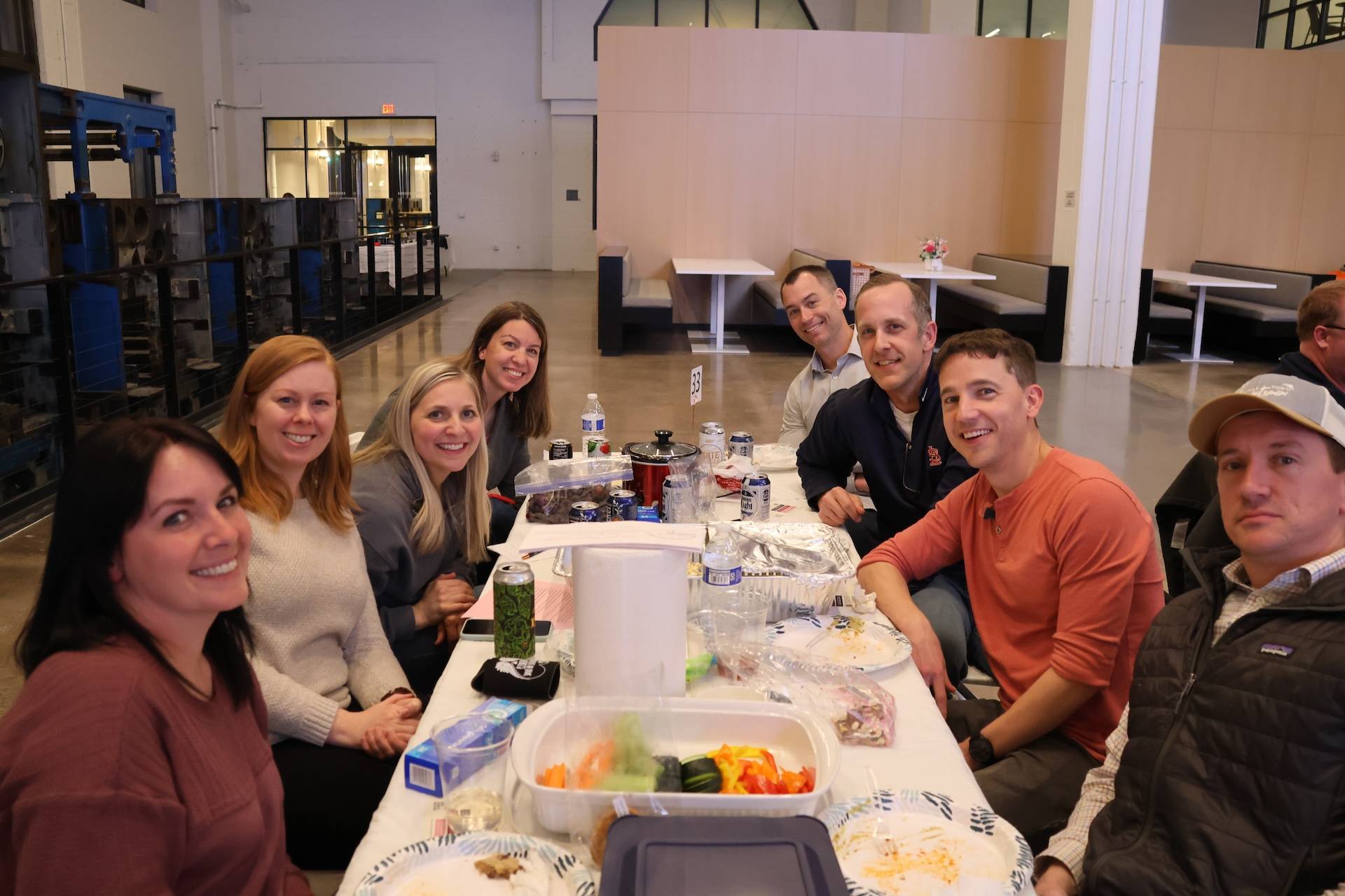 Nine adults seated around a long table indoors, smiling and sharing food, drinks, and plates in a casual cafeteria-style space.