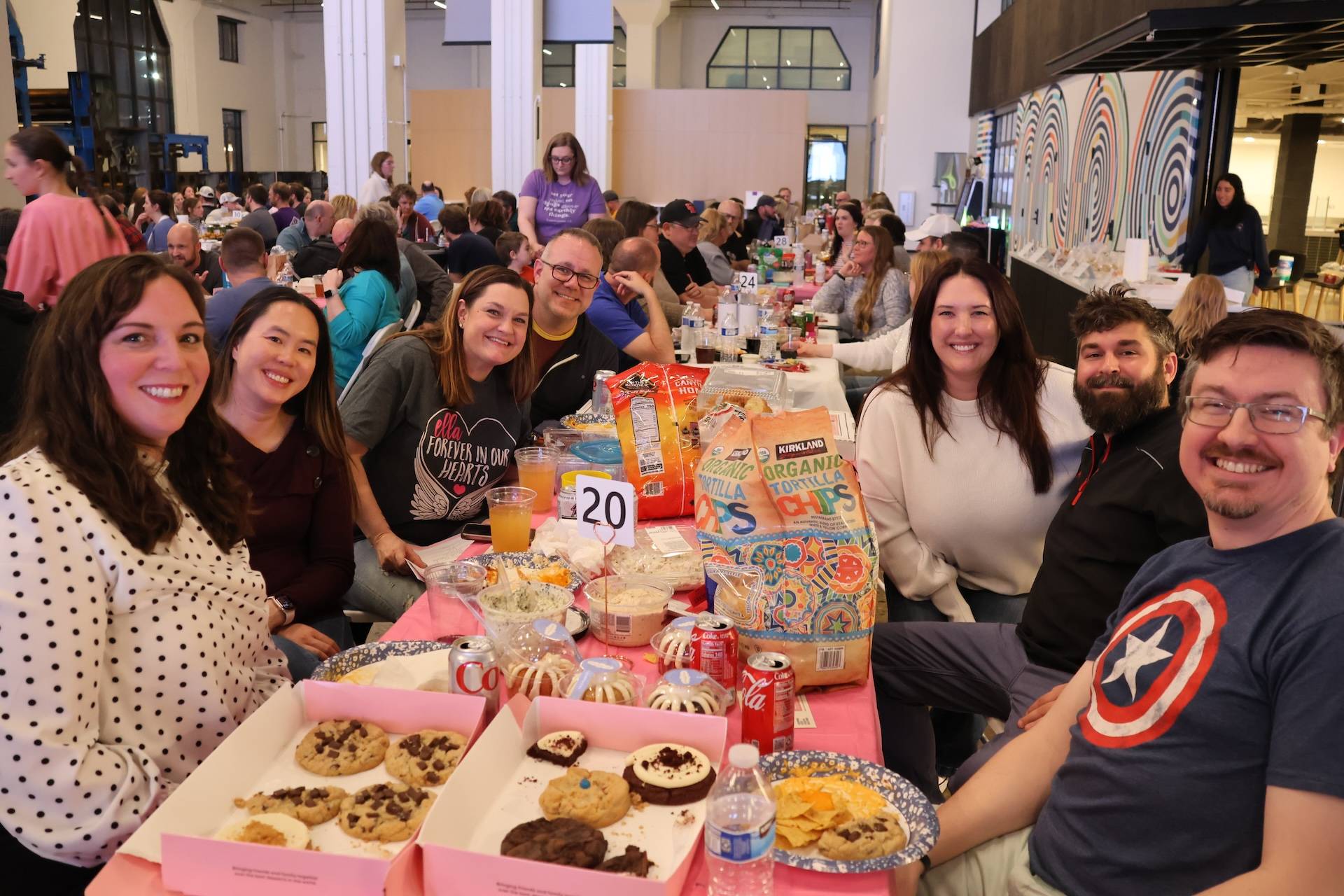 A group of people smiling at a crowded indoor event, seated around a table piled with cookies, chips, drinks, and snacks.
