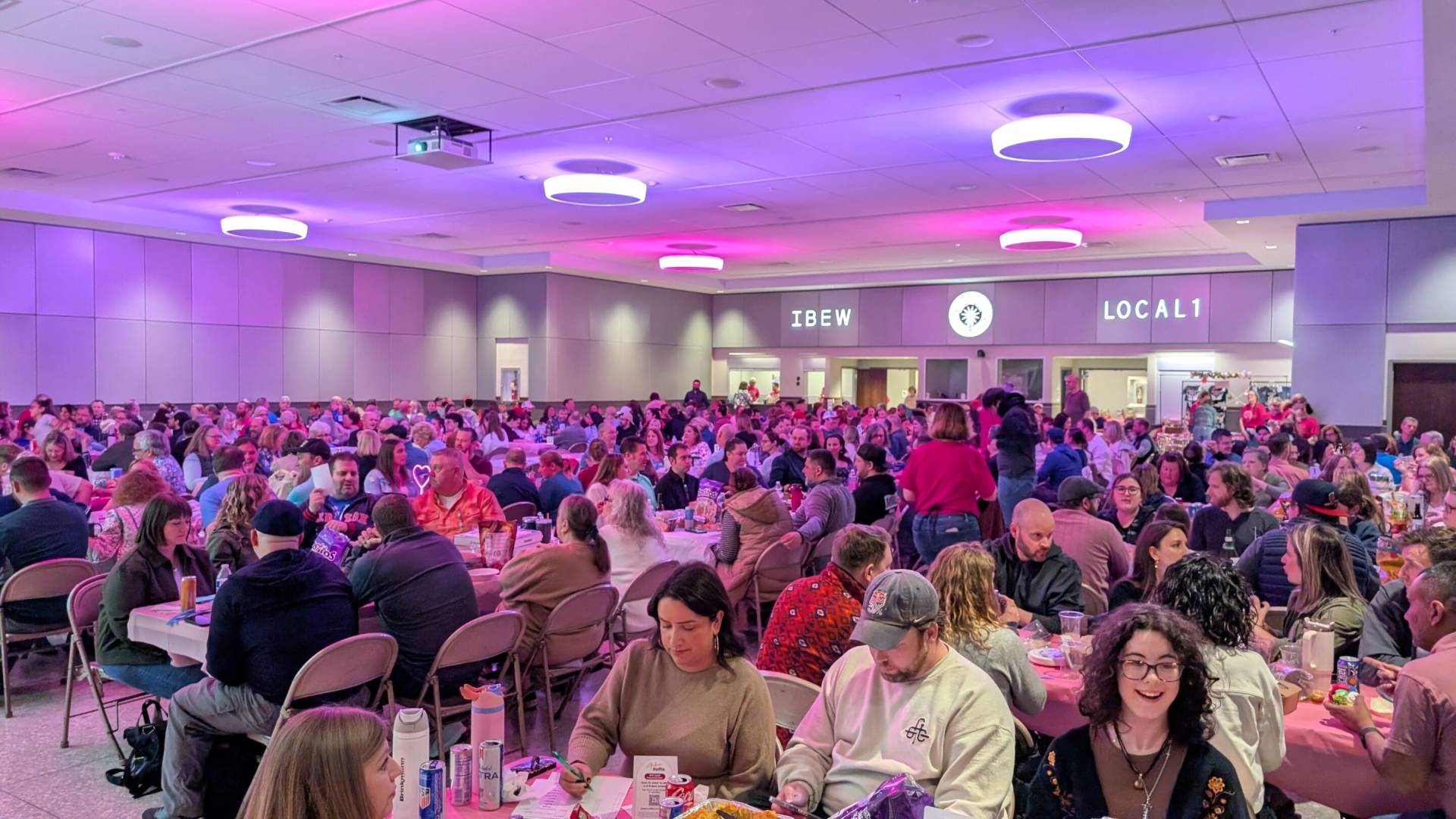 Large banquet hall filled with many people seated at round tables under purple-pink lighting, signs reading IBEW and LOCAL 1 on back wall.