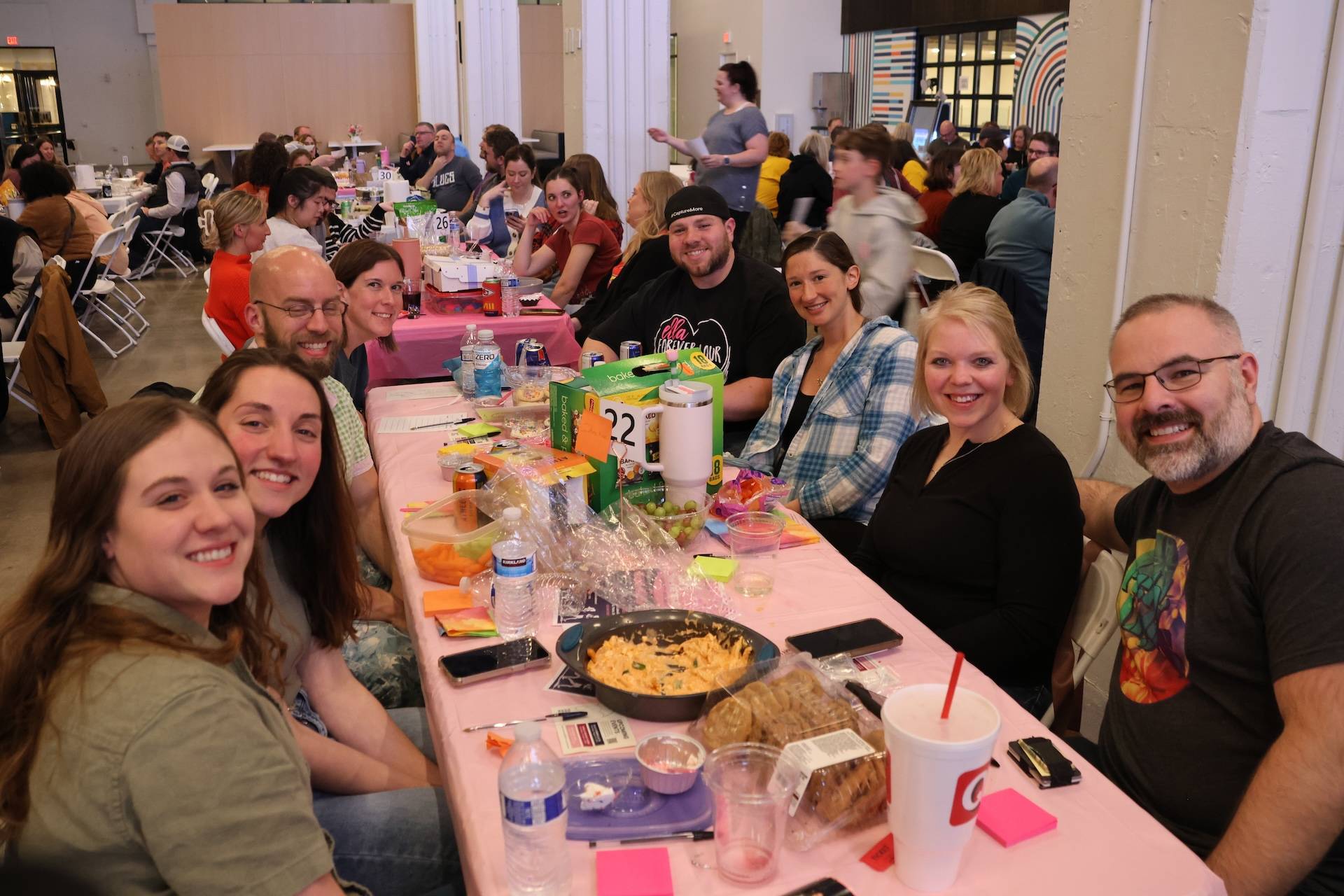Smiling group of adults seated at a long table with pink tablecloth, snacks, drinks, and items at a busy indoor event.
