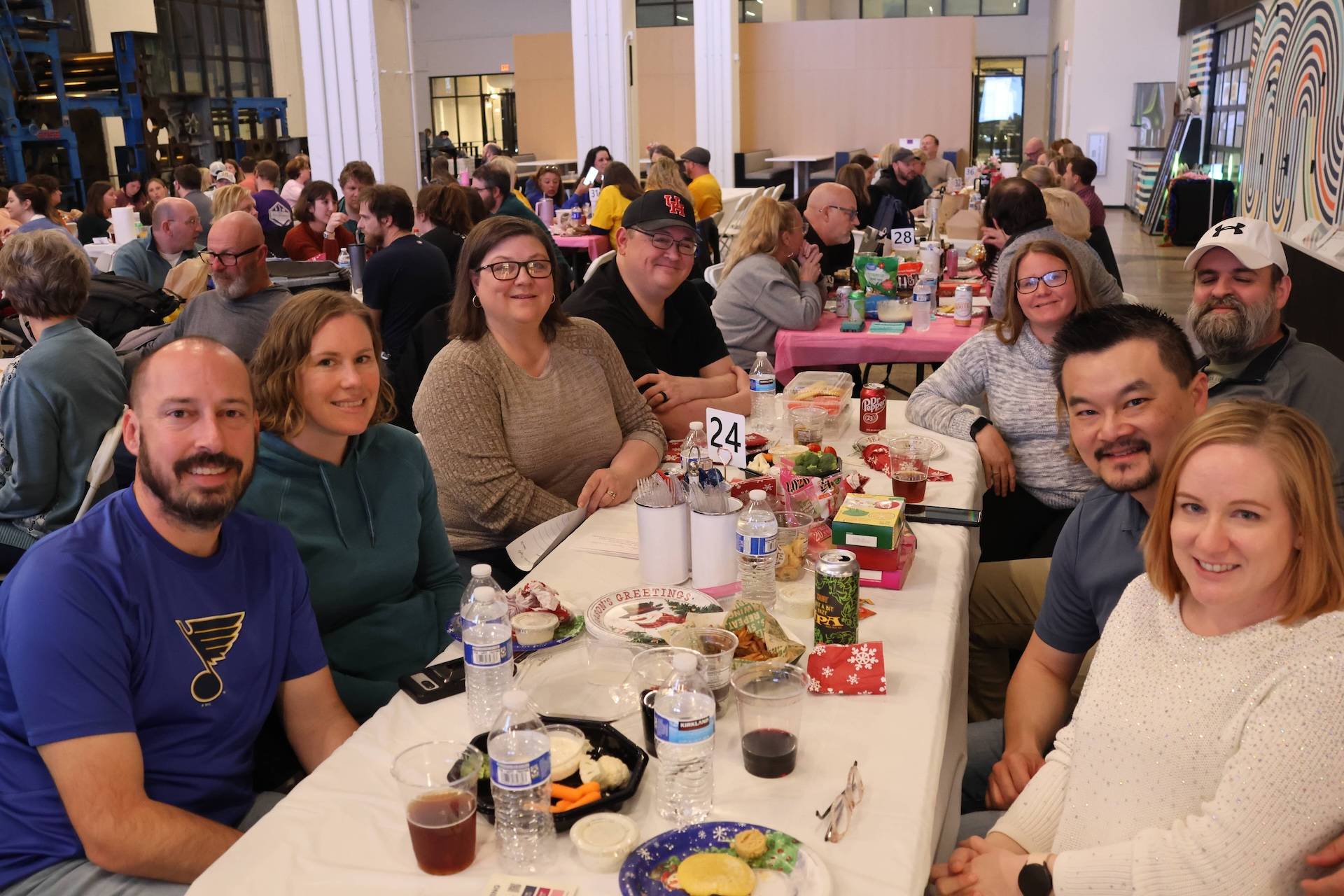 Nine people smiling around a long table laden with food and drinks, with a 24 table card at a busy indoor event