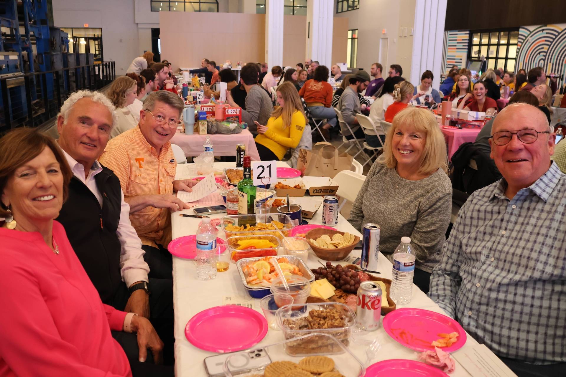 Six smiling adults seated around a table at a crowded gathering with pink plates, snacks, drinks, and a "21" table sign.