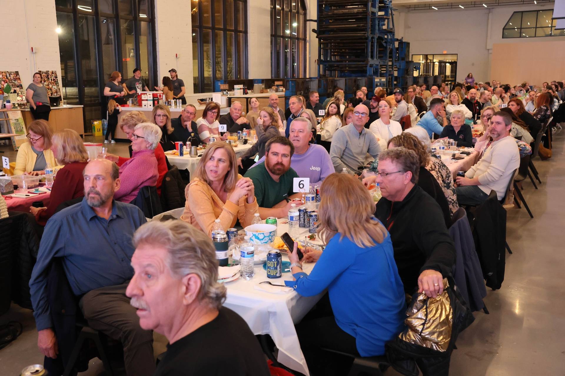 Crowded indoor gathering with dozens seated at numbered banquet tables, many watching toward the front
