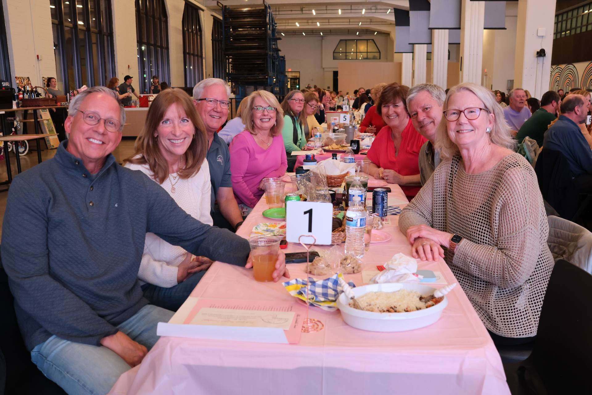 Eight smiling adults seated at a long table with a pink tablecloth, food and a table number 1 in a large indoor event space.