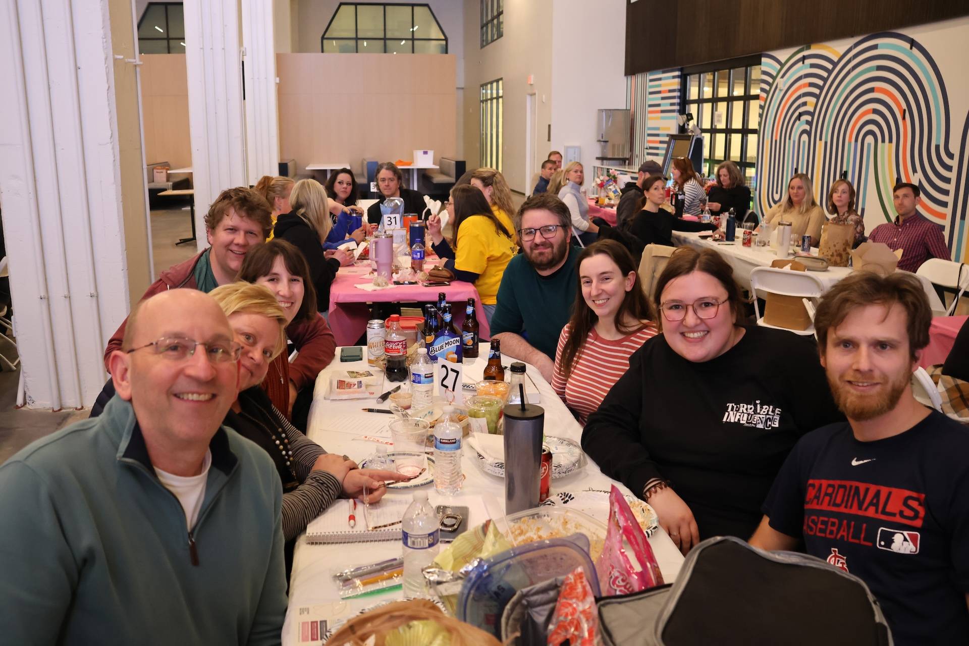 People gathered around a long table at an indoor social event, smiling amid plates, drinks, and table number 27.