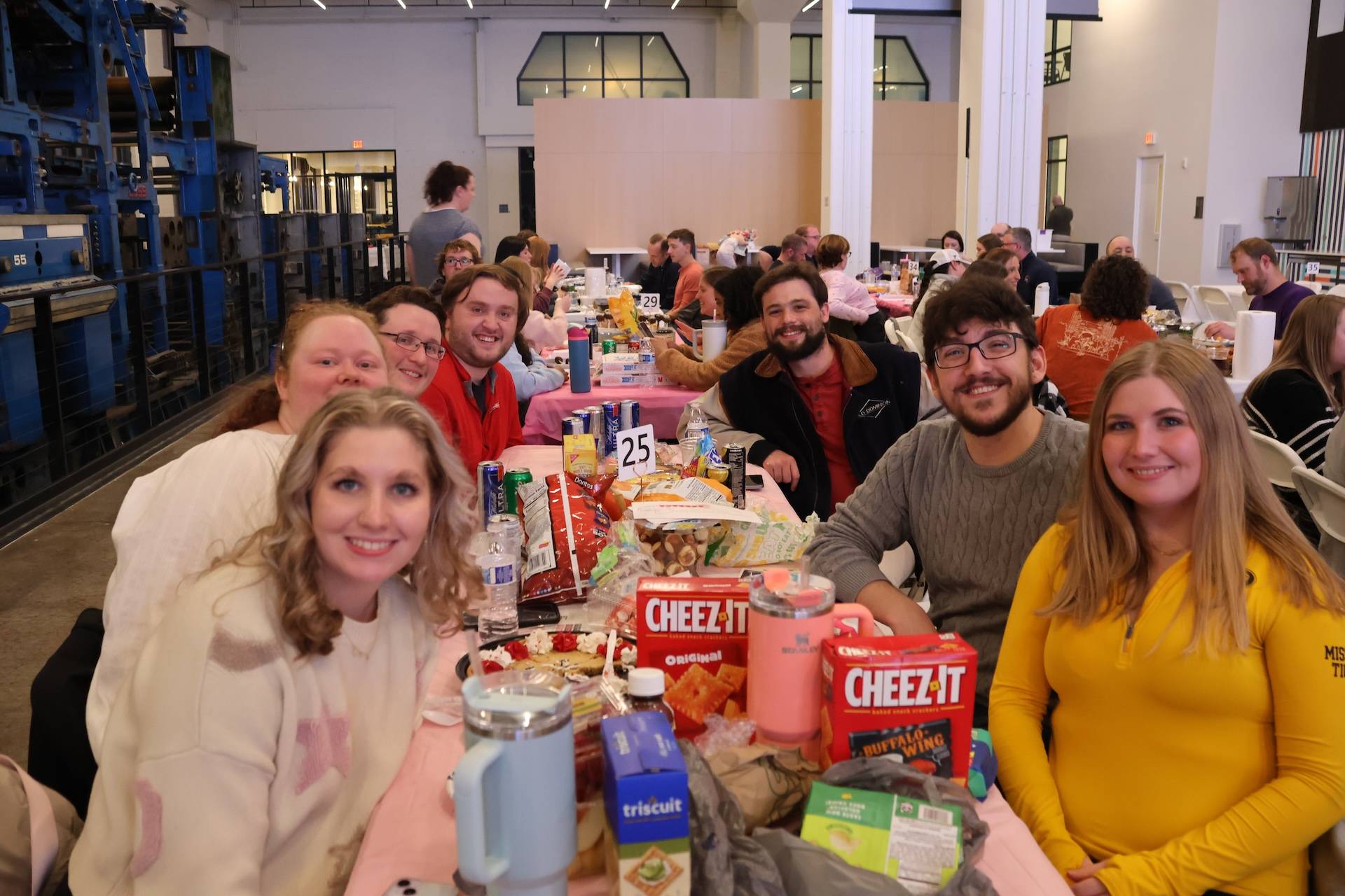 Smiling group at a long indoor banquet table piled with snacks, visible Cheez-It boxes and table number 25.